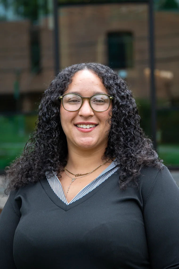 Person with curly hair, wearing glasses and a black top, smiles against an outdoor background with a blurred building.