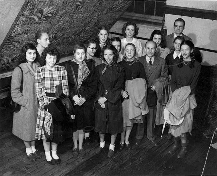 A group of people in mid-20th century attire stand together indoors, with wooden flooring and a decorative wall rug in the background.