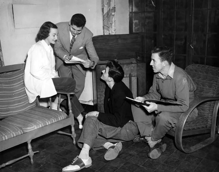 Four people in conversation, two sitting on the floor with notepads and two on a chair and couch. They appear to be discussing papers.