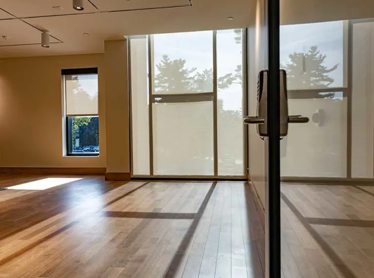 Sunlight streams through large windows onto a wooden floor in an empty room, with shadows casting patterns across the space.