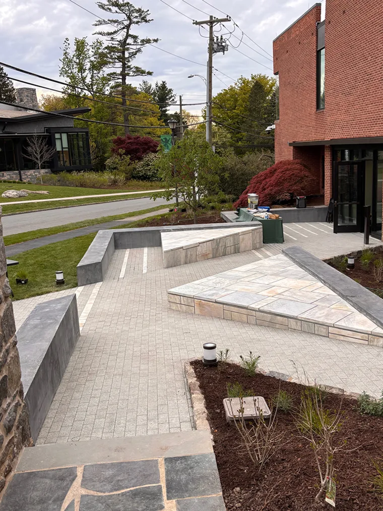 Paved courtyard with geometric patterns, surrounded by brick buildings and greenery. Power lines and trees in the background.