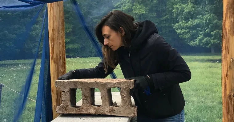 Person inspecting a block structure on an outdoor table, surrounded by trees and a blue canopy.