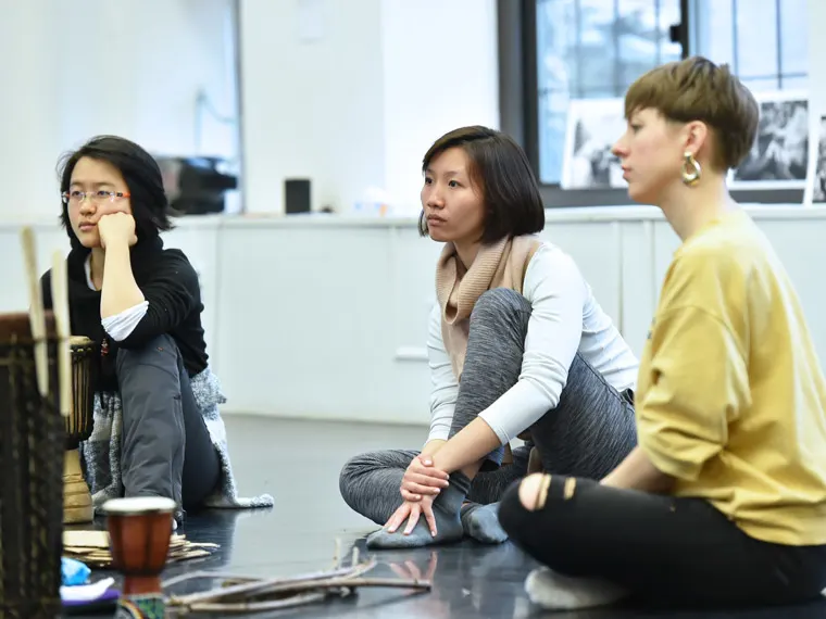Three people sit on the floor attentively during a workshop in a bright room, with musical instruments and photos in the background.