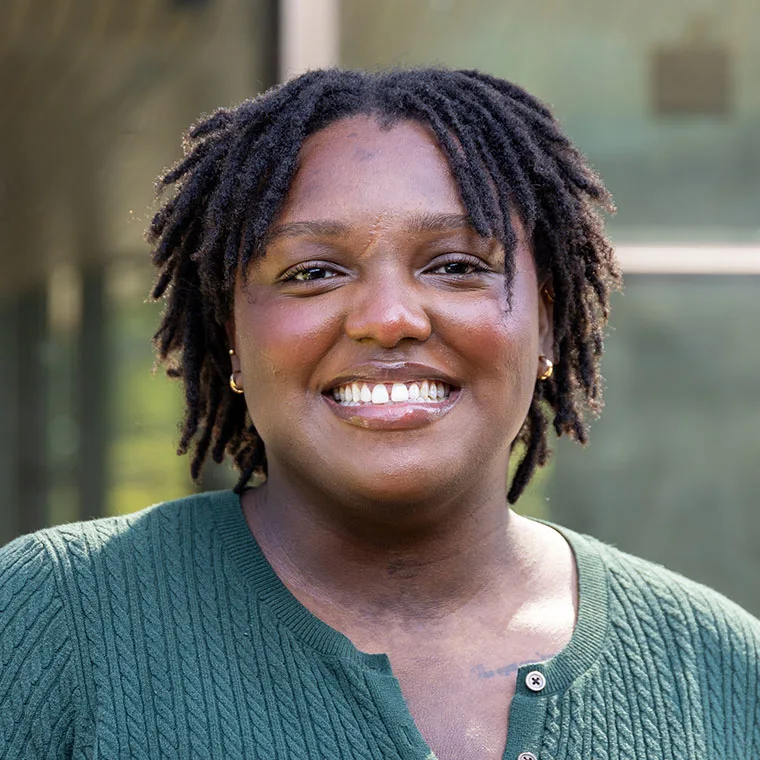 Person with short locs, smiling, wearing a green sweater in front of a blurred outdoor background.