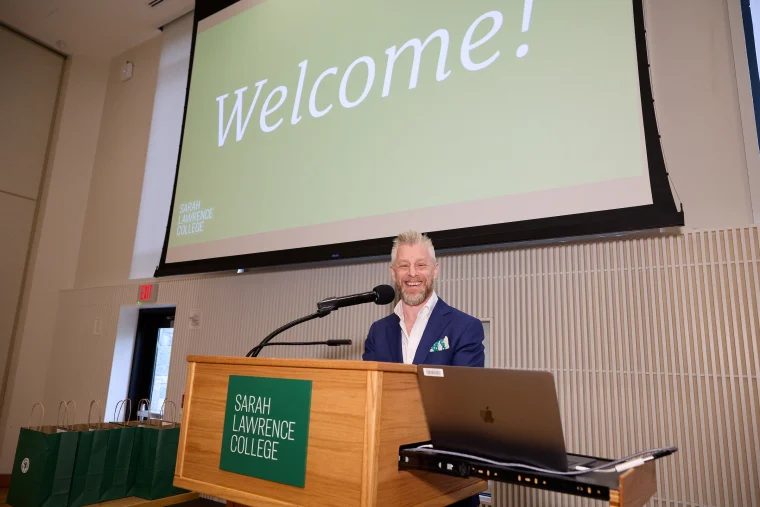 A person speaks at a podium with Sarah Lawrence College signage. A large screen above shows "Welcome!" in green.