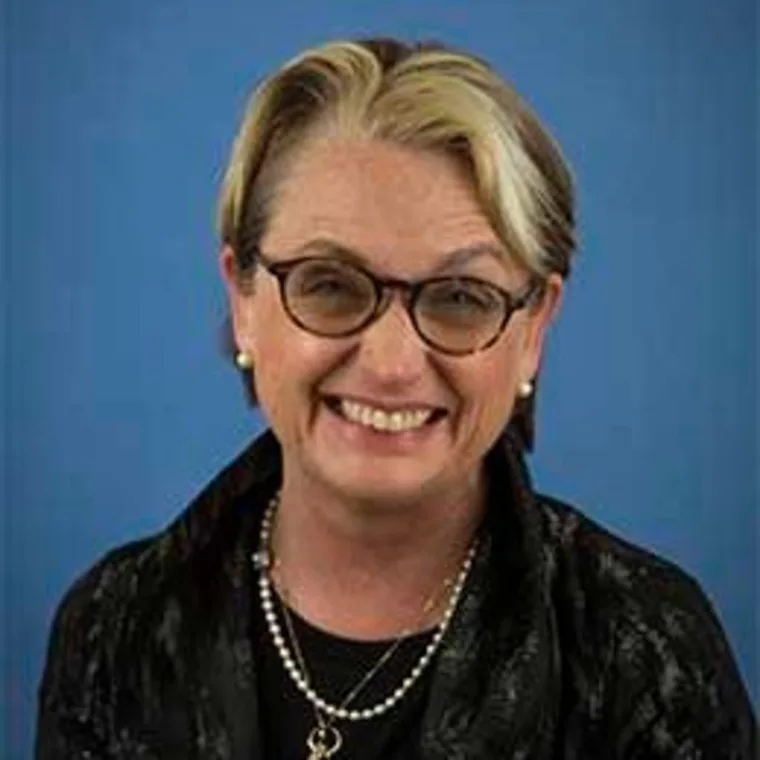 Smiling person with glasses, short hair, wearing a dark jacket, pearl necklace, and earrings against a blue background.