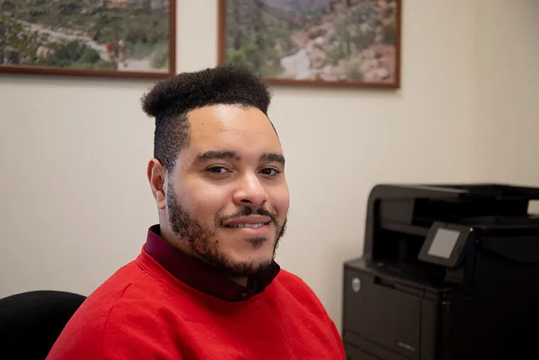 Person in a red sweater smiles in an office with framed landscape photos and a printer in the background.
