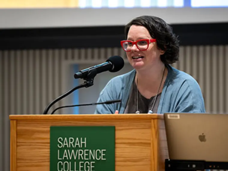 A person with red glasses speaks at a podium labeled Sarah Lawrence College, alongside a microphone and laptop.