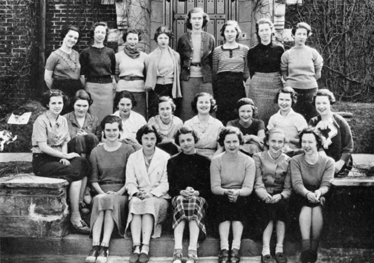 A group of individuals in vintage outfits poses on stone steps outside a building with a large door and brick walls.