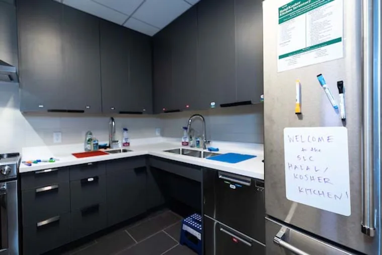 Modern kitchen with dark cabinets, dual sinks, cutting boards, and a refrigerator sign reading "Welcome to the SLC Halal/Kosher Kitchen."