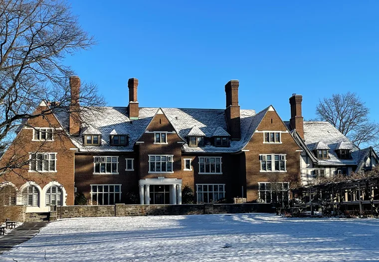 Large brick mansion with multiple chimneys, covered in snow, under a clear blue sky. Trees frame the building, casting some shadows.