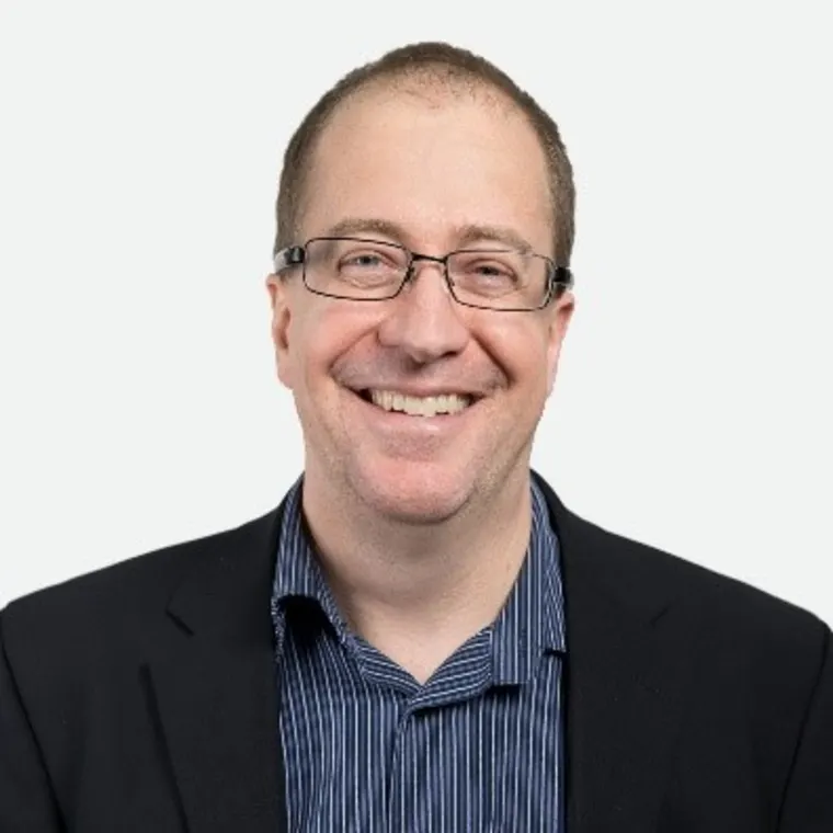Person with glasses smiling, wearing a dark blazer and striped shirt, against a plain background.