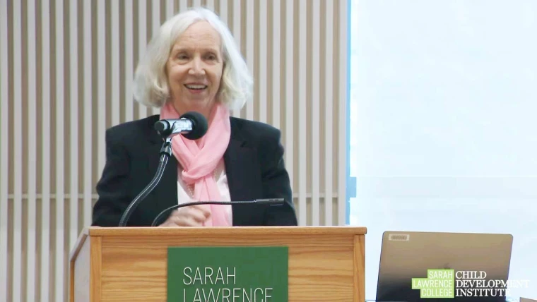 Person speaking at a podium with a microphone, wearing a pink scarf and black jacket. Sarah Lawrence College name is visible on the podium.