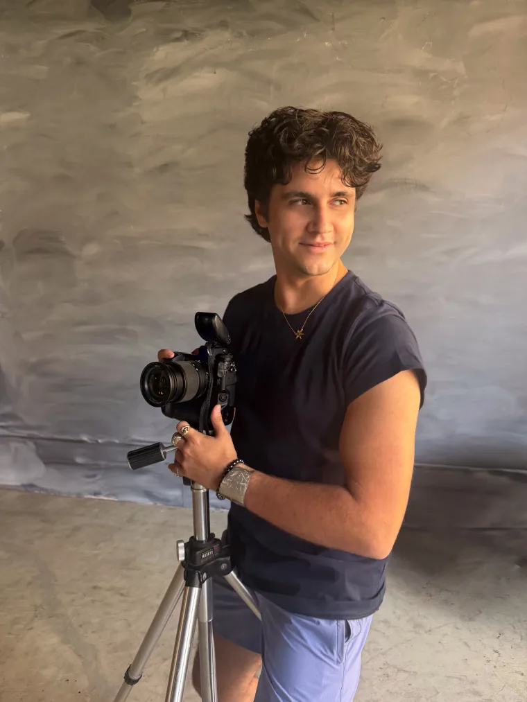 Person holding a camera on a tripod, wearing a dark shirt and light shorts, standing in front of a textured gray backdrop.
