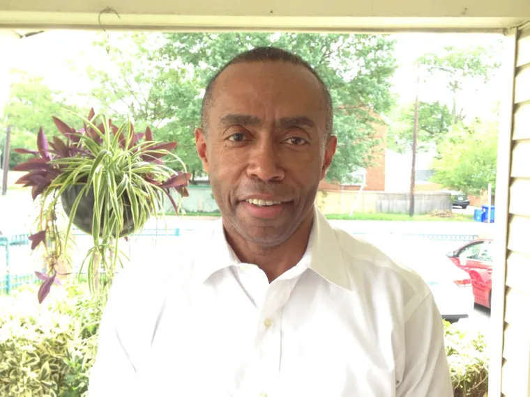 Person in a white shirt smiling, standing in front of a window with greenery and a hanging plant outside.