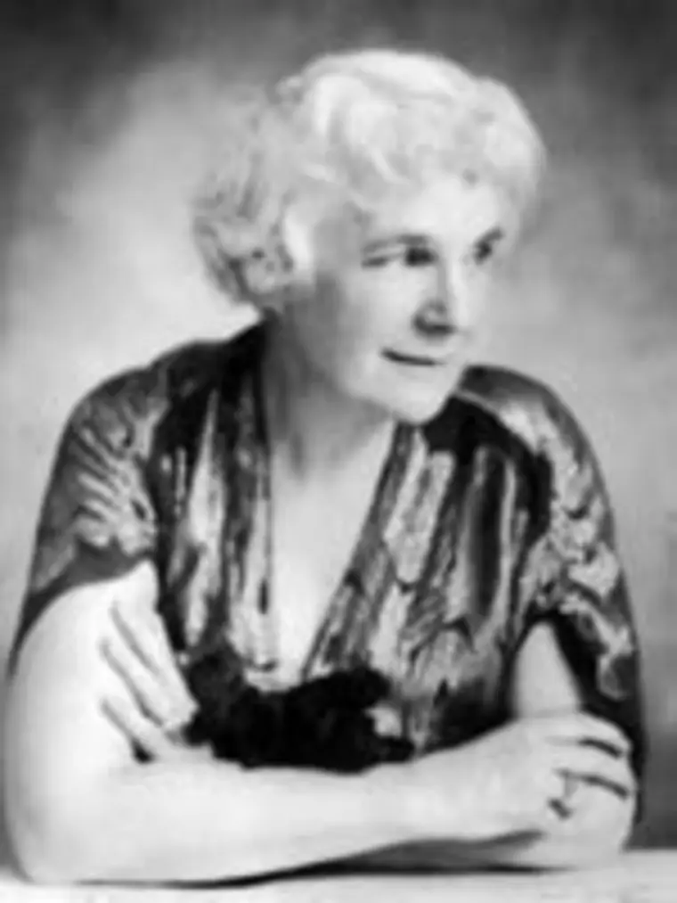 Individual with short white hair, wearing a patterned robe, sits with arms crossed on a table, gazing to the side against a plain background.