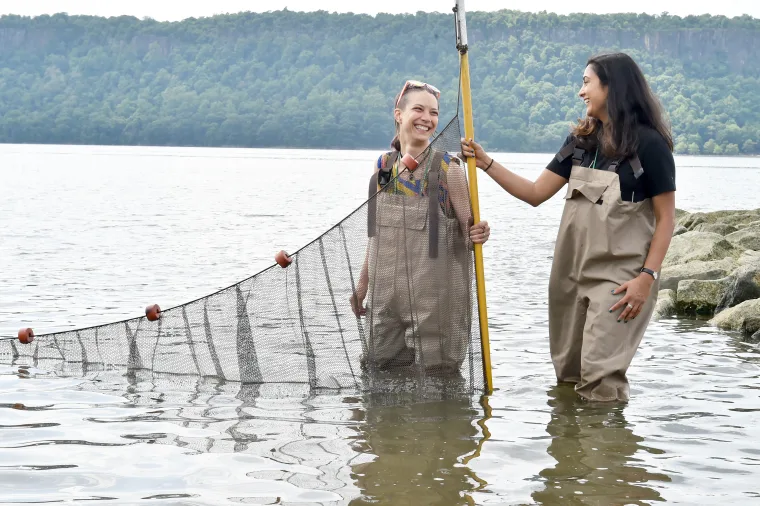 Two people in waders stand in water holding a fishing net. One holds a pole. Trees and hills are visible in the background.