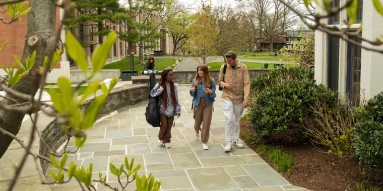 Three people walk on a campus pathway surrounded by trees and a building, engaging in conversation. One person sits on a bench in the background.