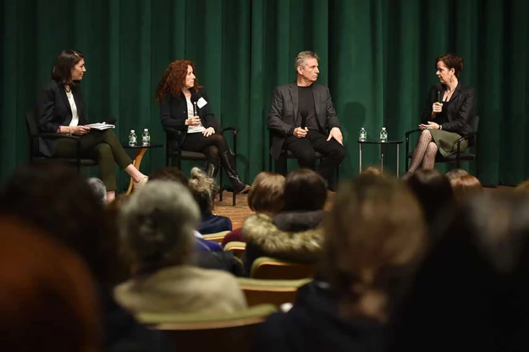 Four individuals seated on a stage for a panel discussion, with a green curtain backdrop and an audience watching.