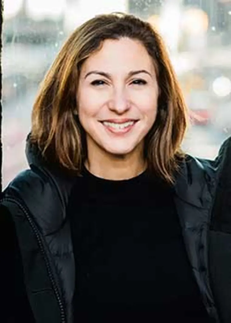 Person with brown hair smiling, wearing a black jacket, blurred outdoor background.