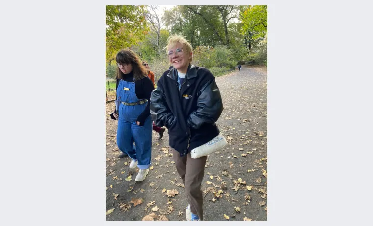 Two people walk on a leaf-covered path in a park, wearing casual jackets. One carries a rolled-up paper under an arm, and trees are visible in the background.