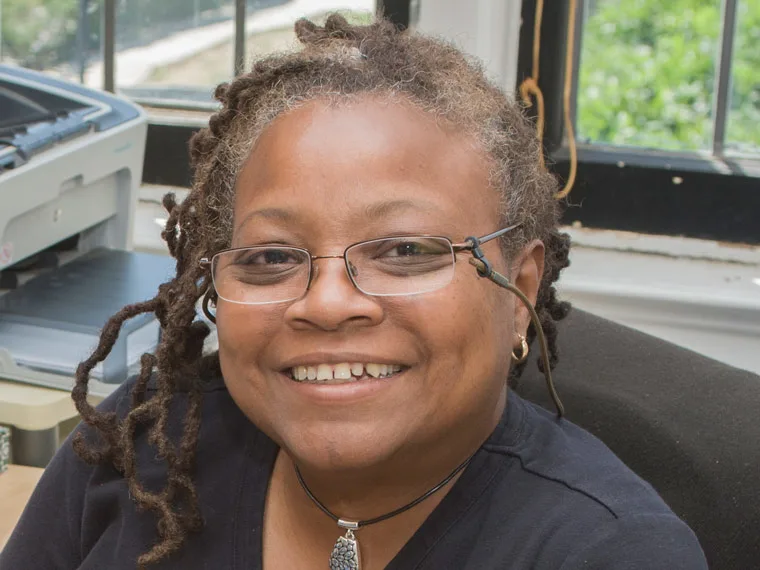 Smiling person with glasses, dreadlocks, and a necklace in an office with a printer in the background.