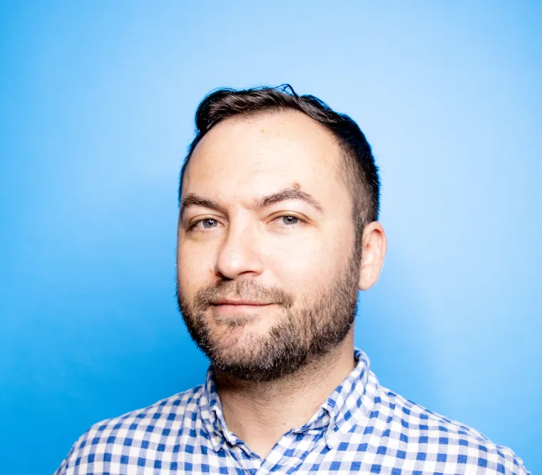 Person with short dark hair and beard, wearing a white and blue checkered shirt, against a bright blue background.