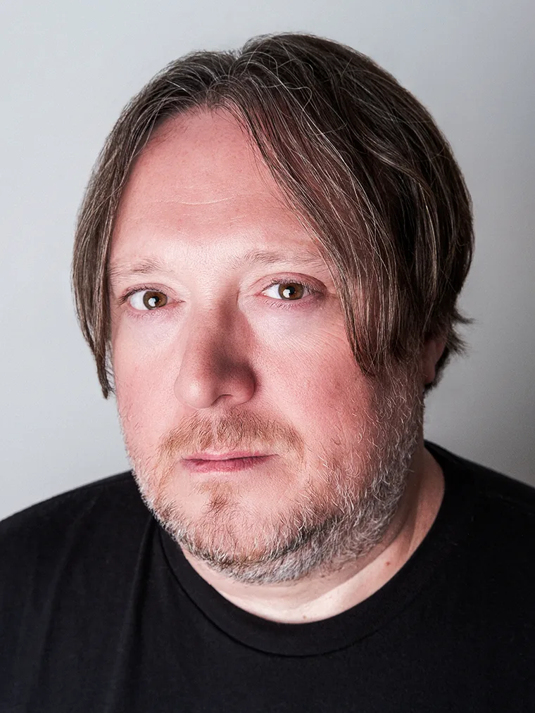Person with shoulder-length hair and a short beard, looking directly at the camera, wearing a black shirt against a light gray background.