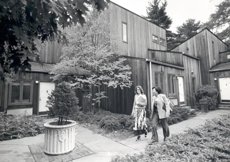 Two people walk on a path beside wooden buildings surrounded by trees and shrubs. A large planter is visible in the foreground.