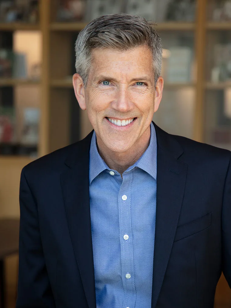 Person smiling in a blue shirt and dark jacket, standing indoors with shelves in the background.