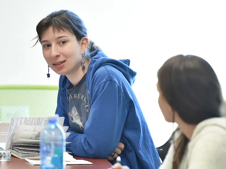 Person in blue hoodie talking at a table, holding papers, with another person blurred in the foreground.