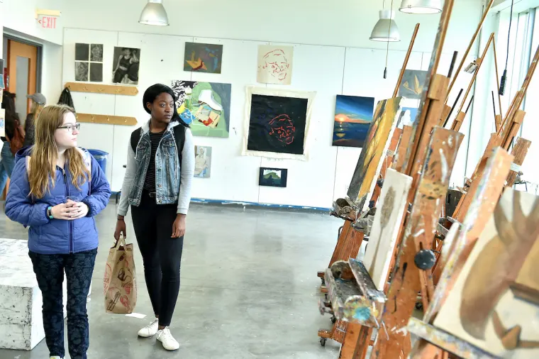 Two individuals in an art studio observe paintings on easels, with various artworks displayed on the walls in the background.