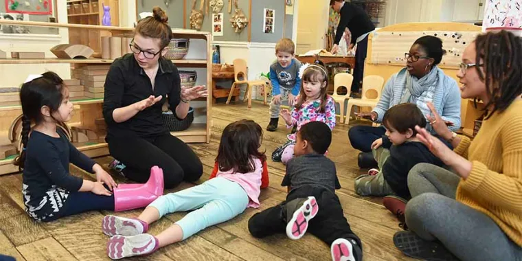 Group of young children and caregivers sitting on the floor in a classroom, engaged in an interactive activity with hand gestures.