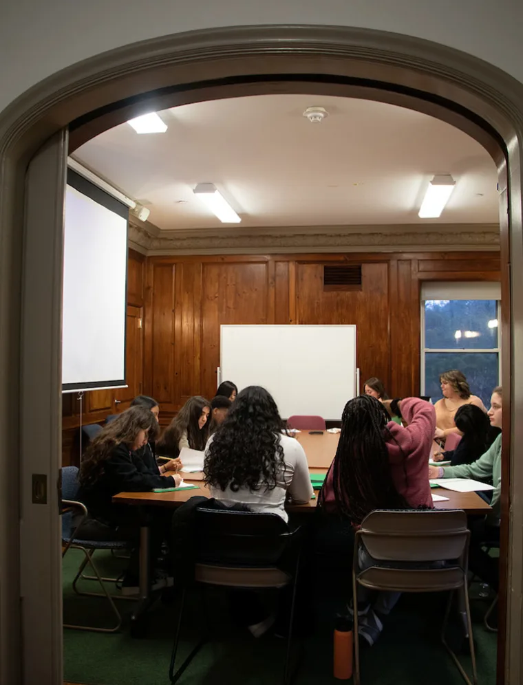 Group of people seated around a table in a wooden-paneled room with a projector screen and whiteboard.