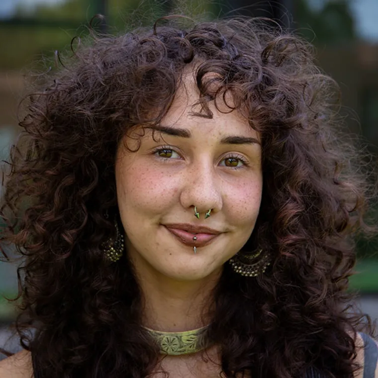 Person with curly hair, freckles, and multiple facial piercings, wearing large earrings and a choker, smiles slightly at the camera.