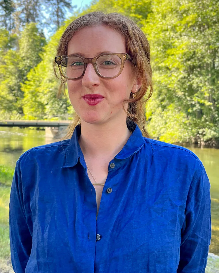 Person with glasses and curly hair in a blue shirt stands outside by a river, surrounded by greenery and trees.