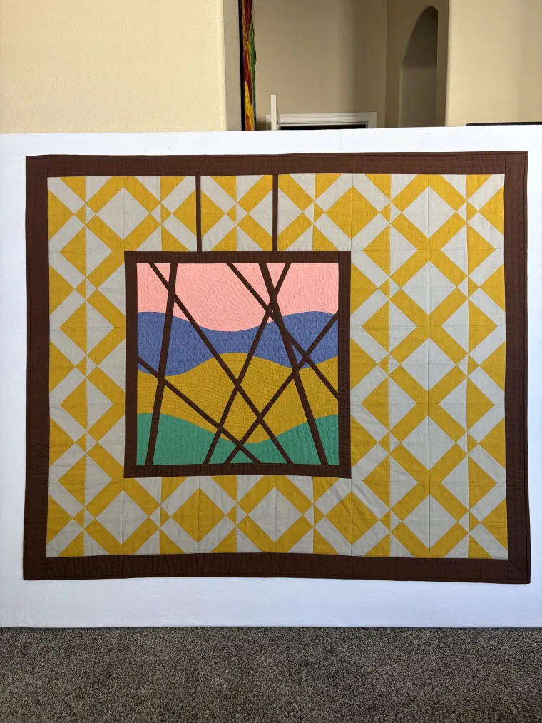 Patchwork quilt with a brown border, featuring a geometric pattern of yellow and beige diamonds, and a central abstract landscape in pink, blue, and green.