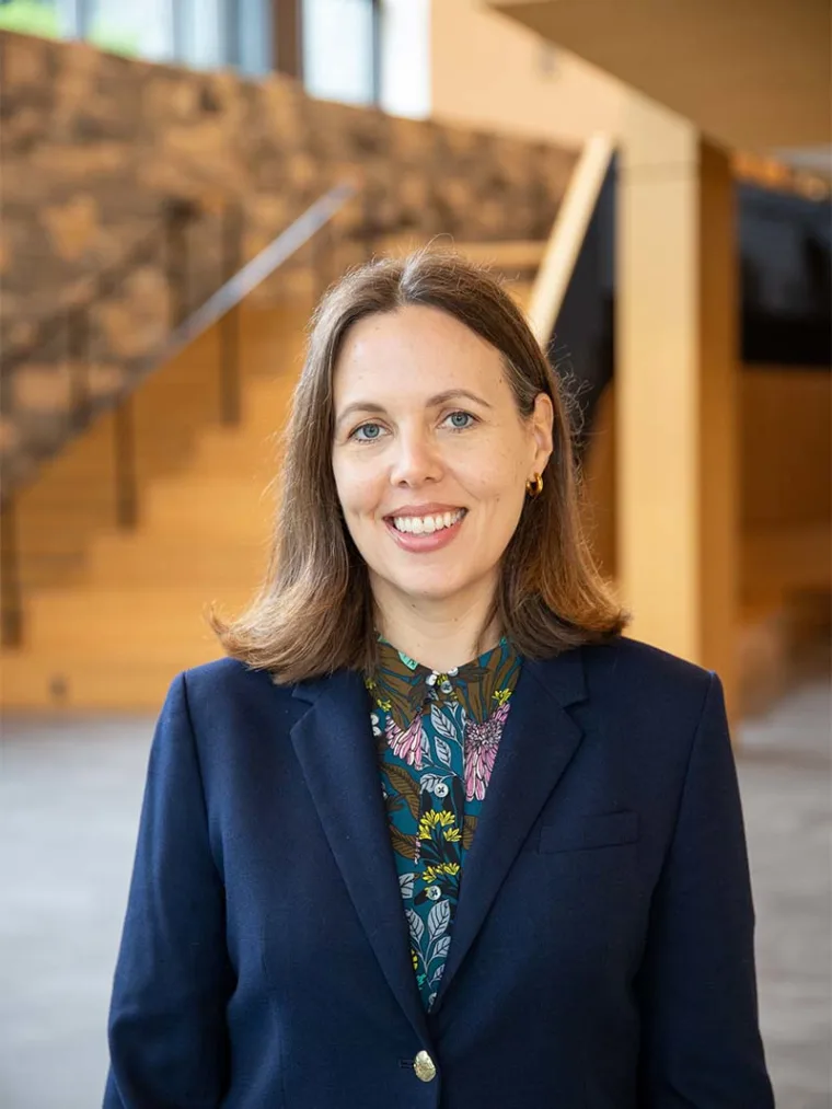 Person smiling wearing a dark blazer and patterned shirt, standing indoors with blurred staircase and wall in the background.
