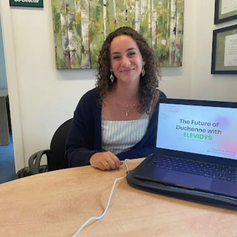 Person sitting at a table with a laptop showing a presentation titled "The Future of Duchenne with Elevidys." Art and certificates on the wall.