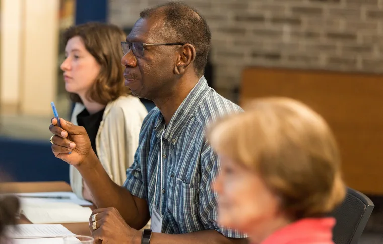 Three people sit at a table, attentively listening and taking notes during a meeting. One holds a pen, and papers are spread on the table.