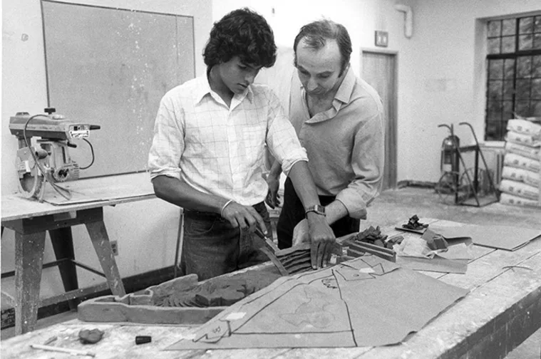 Two people work together in a workshop, examining a paper template on a table surrounded by tools and materials.