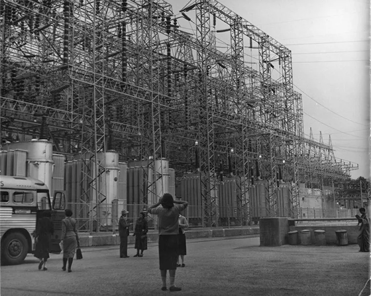 People standing in front of a large industrial power substation with metal structures and transformers, beside a parked bus.