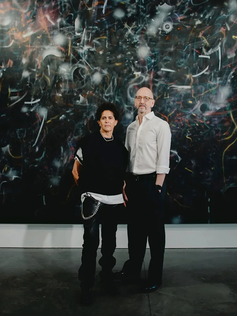 Two individuals stand in front of a large, abstract artwork, one wearing a black outfit with chains, the other in a white shirt.