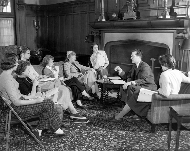 A group of people sit in a semi-circle in a room with wood paneling, engaged in a discussion, with books and papers on their laps.
