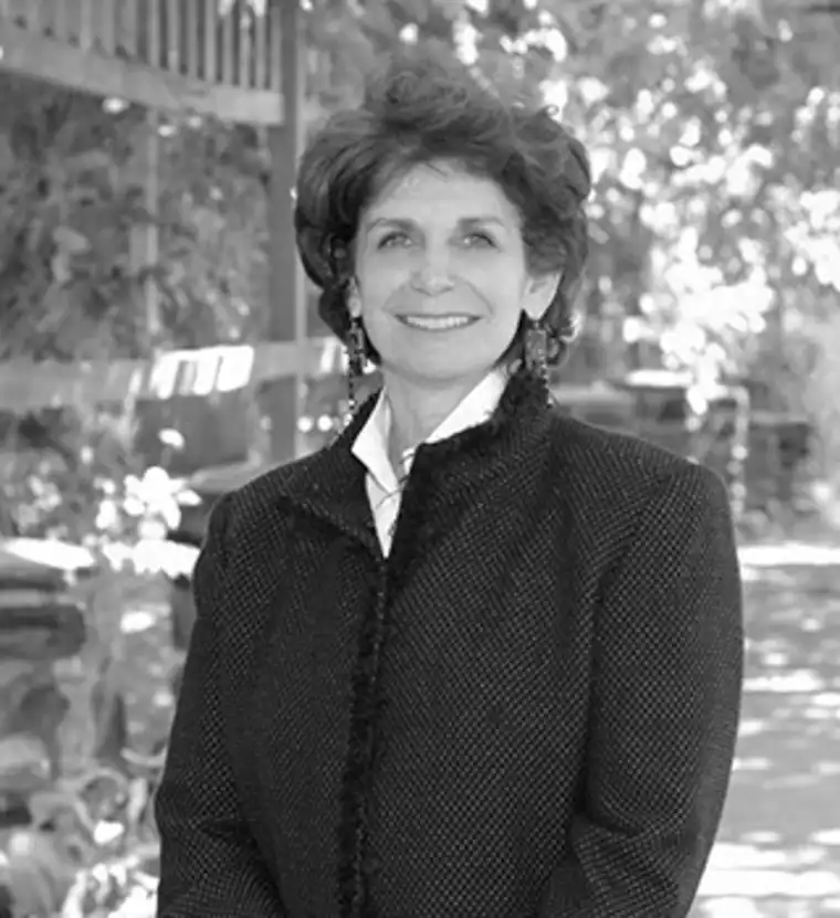 Person in a dark textured jacket stands outdoors in front of foliage, smiling at the camera.