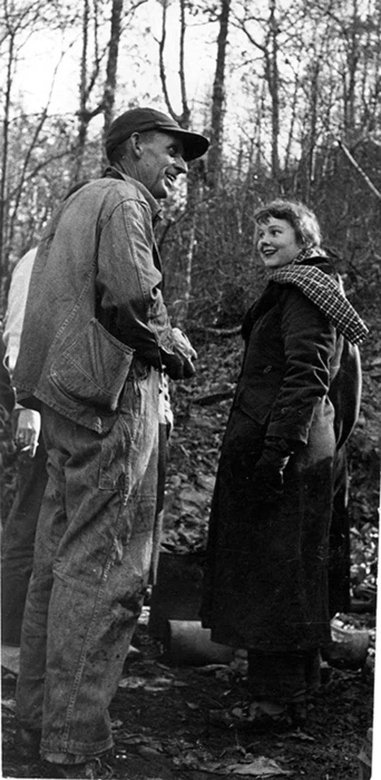 Two people stand outside in a wooded area, wearing coats and smiling at each other. One wears a cap and the other a scarf.
