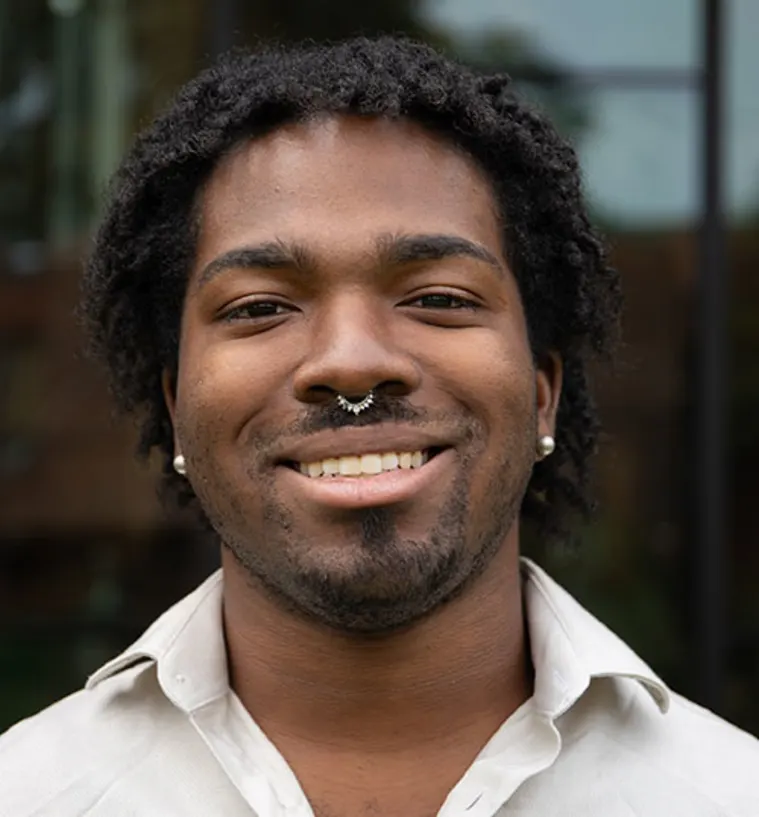 A person smiling, wearing a white shirt, ear piercings, and a septum ring. The background is blurred, showing a building with windows.