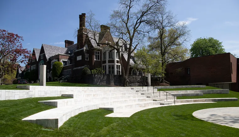 Historic brick building with tall chimneys surrounded by trees and manicured lawns, featuring modern tiered seating in the foreground.