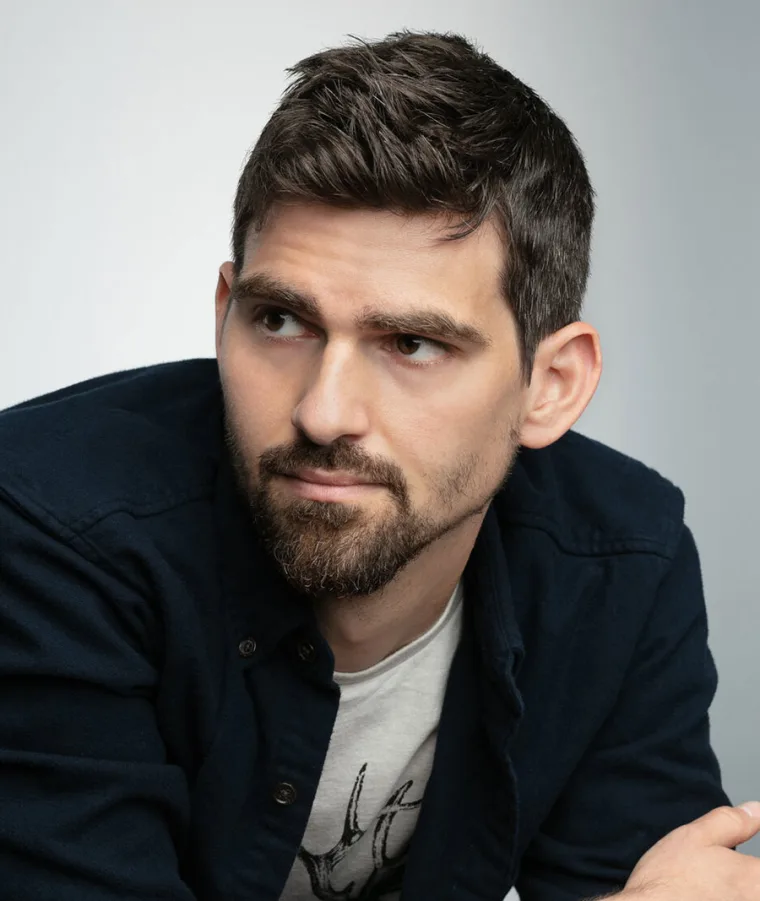 Person with short dark hair and a beard, looking to the side against a gray background, wearing a dark jacket over a graphic T-shirt.