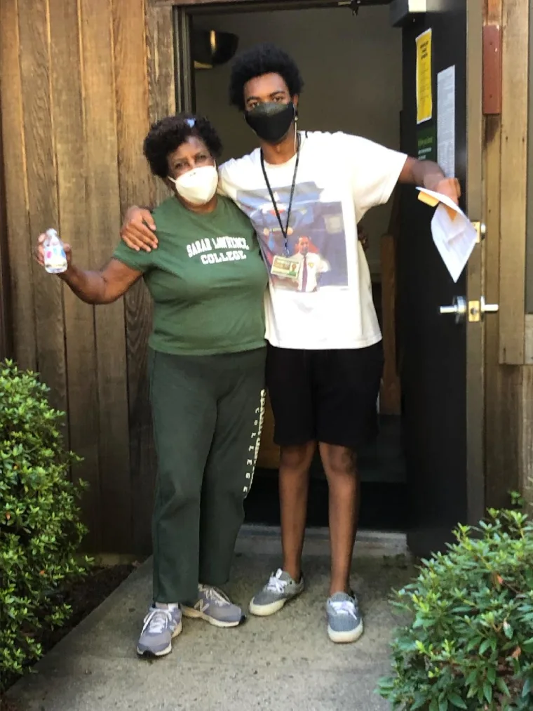 Two people wearing masks stand in a doorway, embracing. One holds a drink, the other holds papers. They are surrounded by greenery.